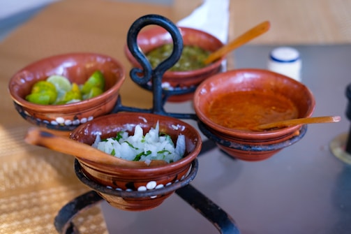 Four small clay bowls are placed on a wrought iron stand. Each bowl contains different ingredients: one has lime wedges, another contains chopped onions and cilantro, a third is filled with a green sauce, and the fourth with a red sauce. Wooden spoons rest in a couple of the bowls. The table underneath appears to be glass.