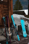 Several pairs of skis and a snowboard rest against a wooden cabin wall outside. The gear is arranged near a stone surface, with a glimpse of snow and a tree in the background, indicating a winter setting.