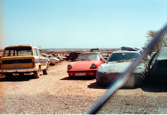 A collection of old and possibly abandoned cars in an open, dusty lot. A red sports car is centrally positioned, partly covered in a layer of dust, surrounded by other vehicles, some of which are covered with tarps. The sky is clear and blue, indicating a sunny day.