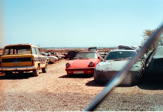 A collection of old and possibly abandoned cars in an open, dusty lot. A red sports car is centrally positioned, partly covered in a layer of dust, surrounded by other vehicles, some of which are covered with tarps. The sky is clear and blue, indicating a sunny day.