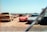 A collection of old and possibly abandoned cars in an open, dusty lot. A red sports car is centrally positioned, partly covered in a layer of dust, surrounded by other vehicles, some of which are covered with tarps. The sky is clear and blue, indicating a sunny day.