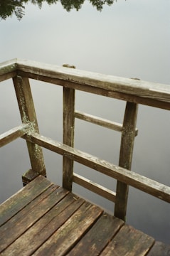 A wooden deck with a rustic, weathered look overhangs calm water. The surface of the water reflects a blurred image of trees, creating a serene and peaceful atmosphere.
