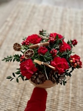 A bouquet of red carnations with green leaves and various decorative elements such as dried citrus slices, pine cones, and cinnamon sticks. The bouquet is held by someone wearing a red sleeve, against a textured neutral background.