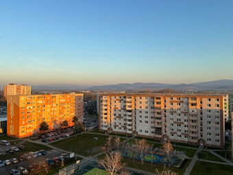 Two apartment buildings with multiple stories are bathed in the golden light of sunset. Cars are parked in the surrounding area, and a playground with a circular design is visible in the foreground. The scene includes open green spaces and trees without leaves. A distant view of mountains can be seen under a clear sky.