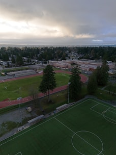 Aerial view of a sports complex surrounded by a residential area. The complex includes a football field with a red running track around it, a soccer field with marked lines, and trees scattered across the area. Beyond the complex is a large building, possibly a school, with parking lots and additional trees visible. The sky above is cloudy, suggesting an overcast day.