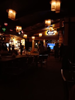 A dimly lit interior of a restaurant or bar with warm ambient lighting. Hanging lanterns cast a soft glow, and there are several tables with chairs. People are casually gathered, suggesting a relaxed and social atmosphere. A neon sign reading 'Brittos' is visible in the background.