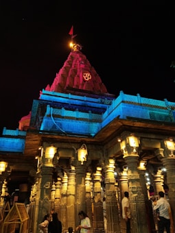 A beautifully lit temple with intricate architectural details is illuminated in vibrant colors of blue and red. The structure features numerous decorative columns and a towering spire with an 'Om' symbol. People are seen gathered around, adding a sense of human activity to the scene.