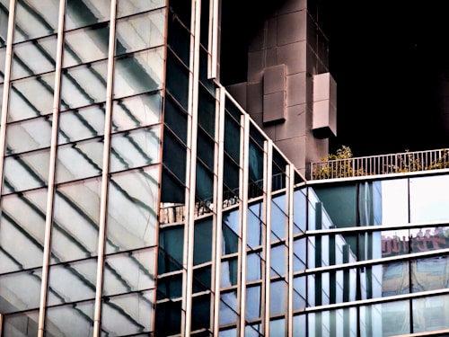 The image features a close-up view of a modern building with a glass facade. The glass panels reflect the surroundings creating a pattern of diagonal lines. A dark structure juts out from the building, adding contrast to the reflective glass. There is a small balcony with some greenery visible on the upper right.
