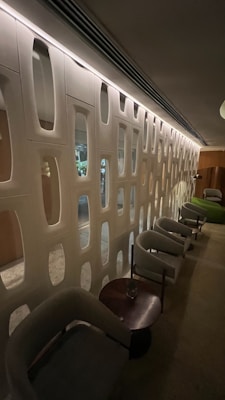 A modern lounge area featuring a series of light-colored modular chairs and small round tables aligned along a uniquely designed wall with alternating vertical and horizontal rectangular cut-outs. Soft ambient lighting illuminates the space, creating a cozy and inviting atmosphere.