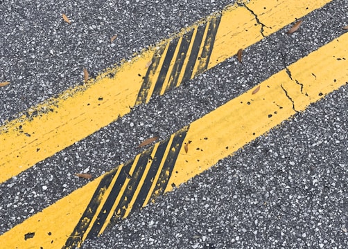 Two parallel yellow lines on an asphalt road with cracks and leaves scattered on the surface.