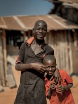 Two boys are standing close together, one taller and one shorter, smiling and posing playfully. They are in front of a rustic building with a corrugated metal roof. The taller boy is wearing a dark shirt, while the shorter is dressed in a bright red shirt.