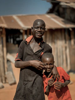 Two boys are standing close together, one taller and one shorter, smiling and posing playfully. They are in front of a rustic building with a corrugated metal roof. The taller boy is wearing a dark shirt, while the shorter is dressed in a bright red shirt.