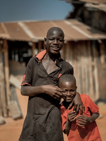 Two boys are standing close together, one taller and one shorter, smiling and posing playfully. They are in front of a rustic building with a corrugated metal roof. The taller boy is wearing a dark shirt, while the shorter is dressed in a bright red shirt.