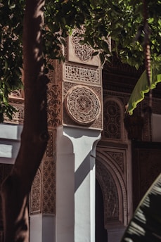 Intricate wall with detailed geometric and floral patterns in a rich architectural setting. The design features arabesque motifs, with Arabic script possibly incorporated. Sunlight filters through adjacent foliage, casting shadows and adding depth.