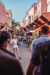 A bustling narrow street lined with colorful buildings and decorative awnings. People meander through the street, shopping and engaging in conversation. Bicycles and market stalls add to the lively atmosphere, capturing a sense of daily life in a vibrant neighborhood.