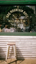 The image features a rustic outdoor scene with a focus on glass signage displaying the words 'BBQ Mountain Boys Burger.' Behind the glass, a reflection suggests a cozy interior with people visible in the background. In the foreground, a simple metal stool stands against a wooden panel wall, adding to the rustic and casual atmosphere.
