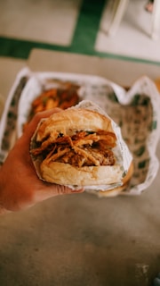A hand holding a hamburger wrapped in paper, featuring crispy fried onions and a beef patty. The background shows a blurred image of fries.