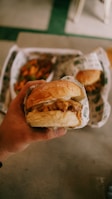 A hand holding a wrapped hamburger with another burger and some fries visible in the background.