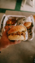 A hand holding a wrapped hamburger with another burger and some fries visible in the background.