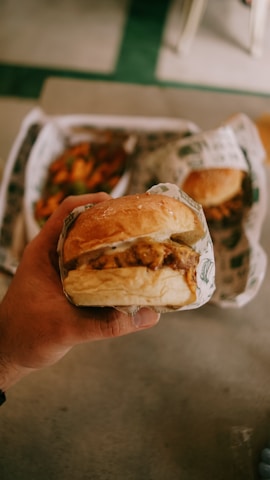 A hand holding a wrapped hamburger with another burger and some fries visible in the background.