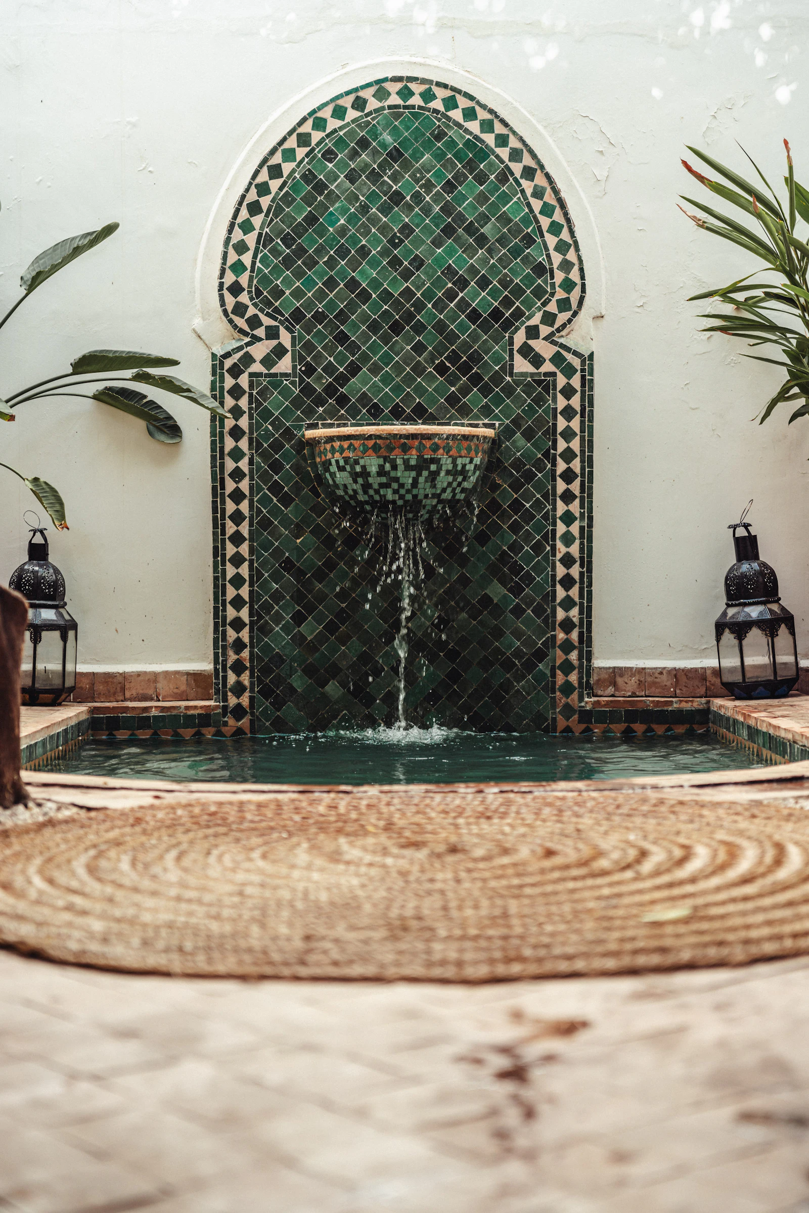 A green zellige-tiled wall fountain trickling into a small pool in a tranquil Moroccan riad, flanked by metal lanterns