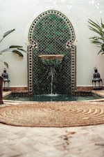 A decorative mosaic tile fountain with intricate green and black patterns is set against a rustic wall. Water gently flows from a spout into a small pool below. Lush green plants flank the fountain, and two Moroccan lanterns are placed symmetrically beside it. The foreground features a woven circular rug on a pale stone floor.