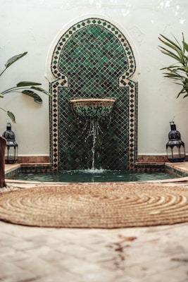 A decorative mosaic tile fountain with intricate green and black patterns is set against a rustic wall. Water gently flows from a spout into a small pool below. Lush green plants flank the fountain, and two Moroccan lanterns are placed symmetrically beside it. The foreground features a woven circular rug on a pale stone floor.