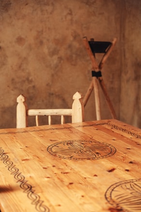 A wooden table featuring intricate circular and linear carvings, set against a textured, rustic wall. A wooden chair with a simple design is partially visible. In the background, a wooden tripod with a black top is placed against the wall.