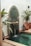A small, decorative fountain against a wall with intricate green mosaic tiles features water pouring from a spout into a small pool. Surrounding the fountain are potted plants and decorative lanterns, adding a touch of greenery and ambiance to the scene.
