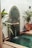 A small, decorative fountain against a wall with intricate green mosaic tiles features water pouring from a spout into a small pool. Surrounding the fountain are potted plants and decorative lanterns, adding a touch of greenery and ambiance to the scene.