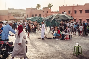 A busy outdoor market scene with a crowd of people engaged in various activities, including walking, chatting, and playing music. The setting features large green umbrellas providing shade, and a backdrop of traditional pink-toned buildings. Motorbikes, stools, and market stalls are scattered throughout the area.
