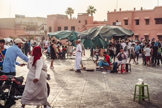 A busy outdoor market scene with a crowd of people engaged in various activities, including walking, chatting, and playing music. The setting features large green umbrellas providing shade, and a backdrop of traditional pink-toned buildings. Motorbikes, stools, and market stalls are scattered throughout the area.