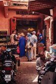 A bustling marketplace scene with several people engaged in conversation near a small shop. The shop has open wooden doors and displays hanging bananas and various produce. There are baskets of fruits and vegetables visible. The narrow street is lined with motorbikes and people appear to be wearing a mix of traditional and casual clothing.
