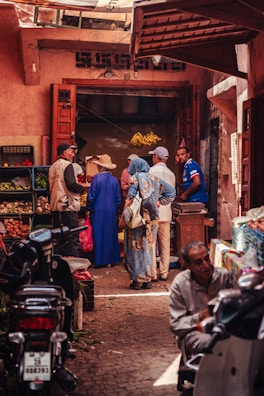 A bustling marketplace scene with several people engaged in conversation near a small shop. The shop has open wooden doors and displays hanging bananas and various produce. There are baskets of fruits and vegetables visible. The narrow street is lined with motorbikes and people appear to be wearing a mix of traditional and casual clothing.