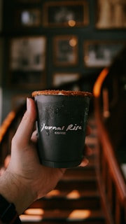 A hand holding a black coffee cup with white text that reads 'Journal Risa Coffee'. The background features a dimly-lit setting with blurred stairs and framed pictures on the wall.