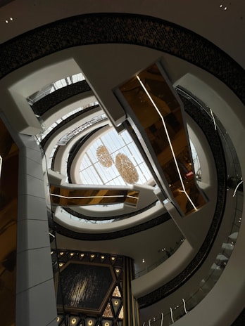 An architectural interior view with multiple layers of curved balconies surrounding a central open space. Above, a skylight ceiling showcases chandeliers, adding a touch of elegance. The design features a mix of white, gold, and dark hues, creating a modern and sophisticated atmosphere.