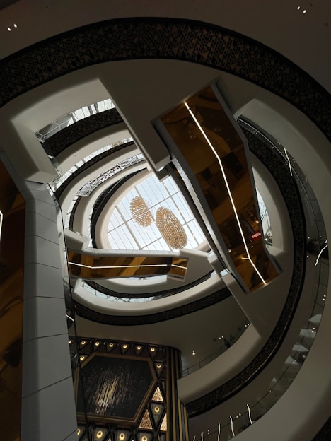 An architectural interior view with multiple layers of curved balconies surrounding a central open space. Above, a skylight ceiling showcases chandeliers, adding a touch of elegance. The design features a mix of white, gold, and dark hues, creating a modern and sophisticated atmosphere.