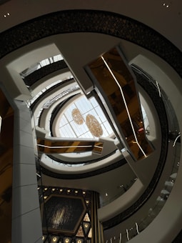 An architectural interior view with multiple layers of curved balconies surrounding a central open space. Above, a skylight ceiling showcases chandeliers, adding a touch of elegance. The design features a mix of white, gold, and dark hues, creating a modern and sophisticated atmosphere.