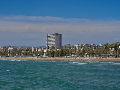 A seaside view features a beach with people enjoying the sunny weather. Palm trees line the shore, providing a tropical atmosphere. In the background, several modern buildings and a tall apartment complex rise against a clear blue sky.
