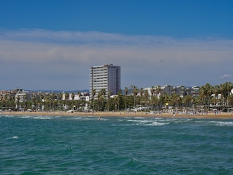 A seaside view features a beach with people enjoying the sunny weather. Palm trees line the shore, providing a tropical atmosphere. In the background, several modern buildings and a tall apartment complex rise against a clear blue sky.