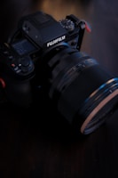A close-up image of a Fujifilm camera placed on a dark surface. The camera features a prominent lens and various control dials visible on top, indicating a professional-grade equipment. The lighting is soft, highlighting the textures and details of the camera's body.