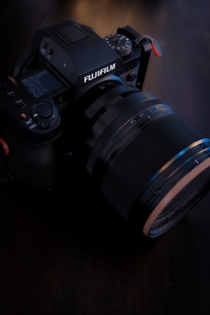 A close-up image of a Fujifilm camera placed on a dark surface. The camera features a prominent lens and various control dials visible on top, indicating a professional-grade equipment. The lighting is soft, highlighting the textures and details of the camera's body.