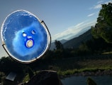 A vibrant blue glass sculpture with circular and oval cutouts is positioned against a picturesque landscape. In the background, rolling hills and mountains stretch into the horizon under a clear sky. Greenery and trees are prominent in the foreground, creating a mix of natural elements.
