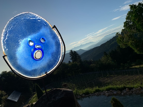 A vibrant blue glass sculpture with circular and oval cutouts is positioned against a picturesque landscape. In the background, rolling hills and mountains stretch into the horizon under a clear sky. Greenery and trees are prominent in the foreground, creating a mix of natural elements.