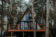 A-frame cabin set in a dense forest surrounded by tall pine trees. The structure features a large glass facade with wooden frames and a spacious wooden deck. Sparse furnishings are visible through the windows, and a parked vehicle is partially hidden behind the trees.