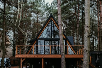 A-frame cabin set in a dense forest surrounded by tall pine trees. The structure features a large glass facade with wooden frames and a spacious wooden deck. Sparse furnishings are visible through the windows, and a parked vehicle is partially hidden behind the trees.