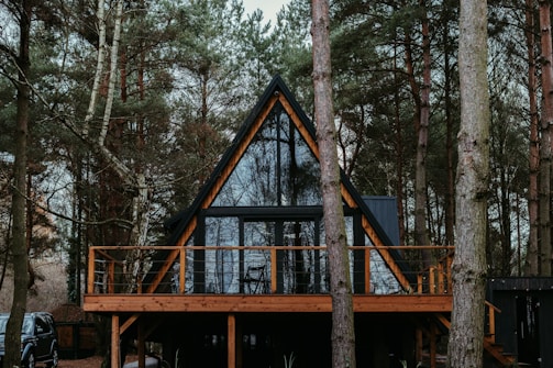 A-frame cabin set in a dense forest surrounded by tall pine trees. The structure features a large glass facade with wooden frames and a spacious wooden deck. Sparse furnishings are visible through the windows, and a parked vehicle is partially hidden behind the trees.