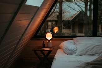 A cozy attic space with a large triangular window showcasing trees and a house in the background. A neatly made bed occupies part of the room, with a soft white pillow and duvet. Beside the bed, a small wooden table holds a modern spherical lamp emitting a warm glow, a small bottle, and a piece of paper. The slanted wooden ceiling adds to the warmth and intimacy of the space.