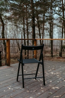 A dark-colored folding chair is placed on a wooden deck surrounded by a wooden railing with cable wires. In the background, a forest with bare trees during the fall or winter season is visible. Dry leaves are scattered on the deck.