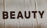 Large, bold letters spelling the word 'BEAUTY' are displayed on a textured surface. The background appears to have a subtle beige tint with visible creases or folds.